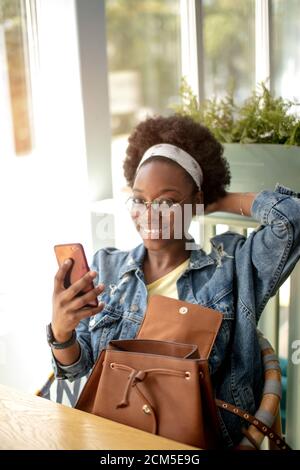 Gut aussehende afrikanische Frauen in Jeans Jacke wirft bei Camera von smart phone Holding Leder Rucksack, wobei selfie, genießt, Freizeit oder Wochenende. Stockfoto