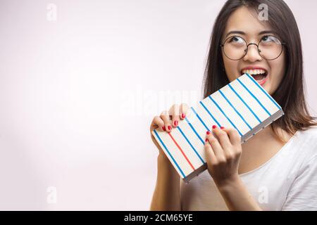 Aufgeregt verspielte asiatische Mädchen mit dunklen Haaren, beißt ein Buch, schaut zur Seite, trägt Brille, Emotionen vor der Prüfung. Stockfoto