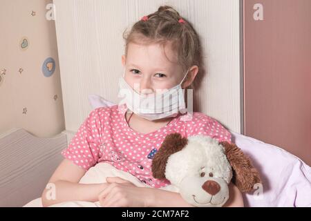 Baby Mädchen trägt Schutzmaske in Quarantäne, liegt im Bett mit einem weichen Spielzeug. Patient im Haus isoliert, um Infektionen zu verhindern. Coronovirus-Konzept. hor Stockfoto