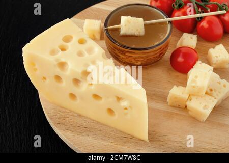 Gelber Maasdam-Käse, dreieckiger Käse mit Löchern, Untertasse mit Honig, Tomatenholz schwarzen dunklen Stein Schiefer Hintergrund Stockfoto