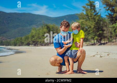 Vater hockt halten Sohn auf offener Hand, spielen vergleichen ihre Handflächen, tragen Gesicht blaue Maske schützen Coronavirus Wandern Natur Sand Meer Strand Stockfoto