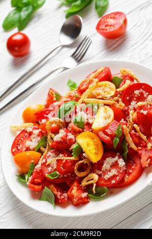 Tomatensalat mit knusprig gebratener Zwiebel, Quinoa und frischem Basilikum auf einem weißen Teller auf einem Holztisch, Nahaufnahme, vertikaler Blick von oben Stockfoto