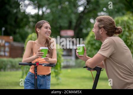 Zwei Personen mit Motorroller reden im Park und haben Kaffee zusammen Stockfoto