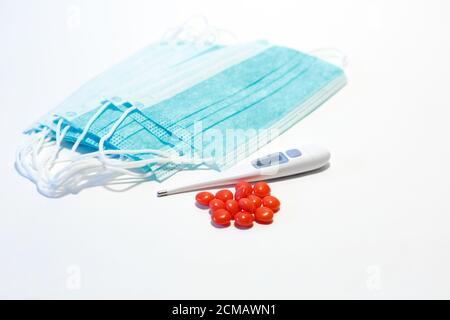 Antivirale Maske zum Schutz vor COVID-19, rote Tabletten, ein elektronisches Thermometer zur Temperaturmessung auf weißem Hintergrund. Ausbruch Des Coronavirus Stockfoto