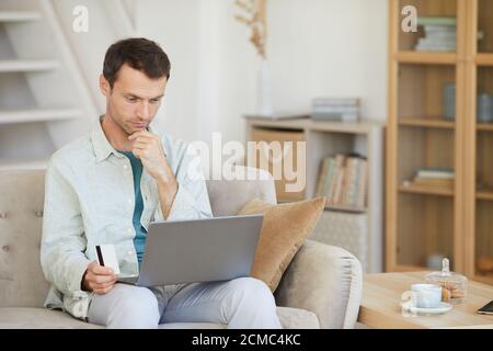 Junger Mann sitzt auf dem Sofa mit Laptop er geht Online mit Kreditkarte zu Hause bezahlen Stockfoto