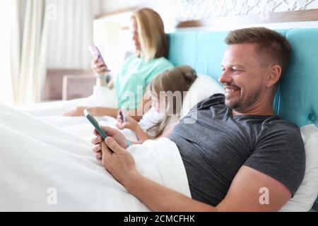 Mama, Papa und Tochter liegen im Bett und schauen sich das Telefon an. Stockfoto