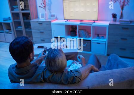 Rückansicht eines jungen Paares, das sich umarmt, während es auf dem Sofa sitzt Popcorn essen und einen Film ansehen Stockfoto
