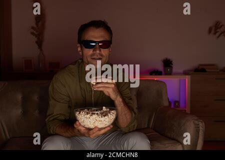 Junger lächelnder Mann in 3D-Brille sitzt auf Sofa essen Popcorn und 3D-Filme zu Hause ansehen Stockfoto