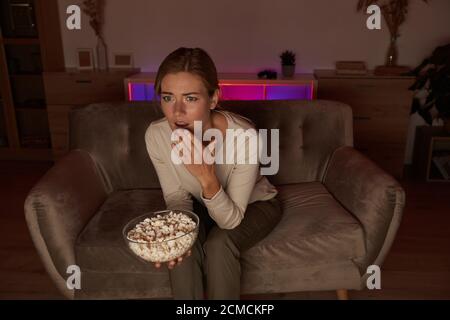 Verängstigte junge Frau auf dem Sofa sitzen und essen Popcorn während Schaue einen unheimlichen Film an Stockfoto