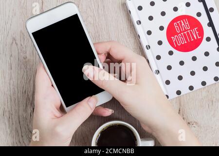 Business- und Bürokonzept - Frau Hände halten weißen smartwhone Neben dem schwarz-weißen Notizbuch mit Punktmuster Und eine Tasse schwarzen Kaffee auf Woo Stockfoto
