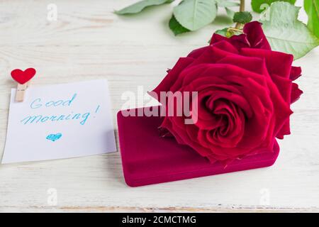 Schmuckschatulle und wunderschöne rote Rose auf Holzhintergrund. Guten Morgen auf weißem Blatt Papier Stockfoto