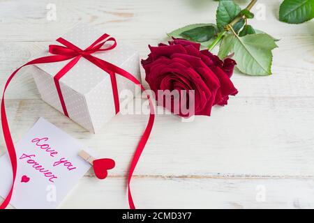 Beigefarbene Geschenkschachtel mit roter Schleife und schönen roten Rosen auf Holzhintergrund. Grußkarte für Urlaub. Stockfoto