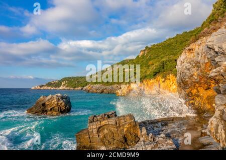 Sommerabend an einem kleinen Strand zwischen den Felsen. Surfspray. Dächer der Hütten zwischen den dichten Wald Stockfoto