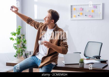 Junger Geschäftsmann, der Selfie auf dem Smartphone hält, während er Tasse davon hält Kaffee Stockfoto