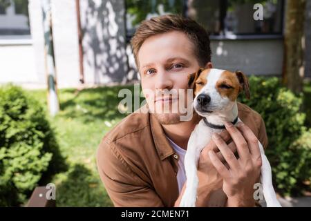 Junger Mann, der die Kamera anschaut, während er Jack russell Terrier kuschelt Hund draußen Stockfoto