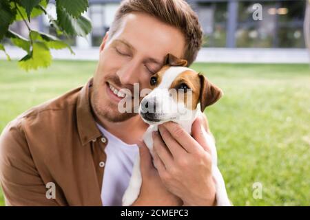 Junger Mann mit geschlossenen Augen, der weißen Jack russell Terrier hält Hund mit braunen Flecken auf dem Kopf Stockfoto