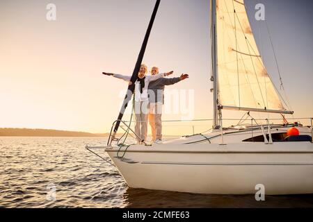Die ganze Länge der glücklichen Senior Paar Stretching Hände heraus gegen den Himmel, genießen Sie erstaunliche Sonnenuntergang und Segeln zusammen, während auf der Seite der Stockfoto