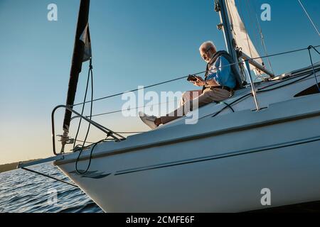 Genießen Sie das Leben. Seitenansicht des älteren Mannes, der auf der Seite des Segelbootes oder der Yacht sitzt, die im ruhigen blauen Meer bei Sonnenuntergang schwimmt und digitale Tablette oder benutzt Stockfoto