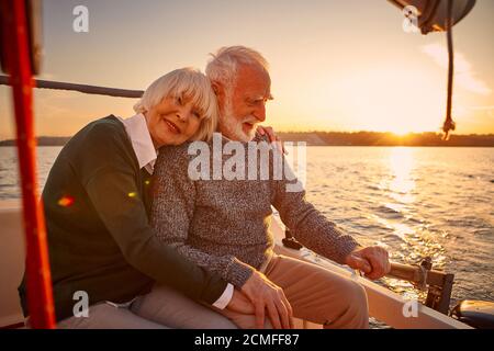 Segeln bei Sonnenuntergang genießen. Glückliches Seniorenpaar, älterer Mann und Frau, die Hände halten, umarmen und entspannen, während sie auf der Seite des Yachtdecks sitzen Stockfoto