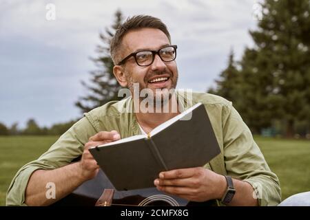 Junger glücklicher Mann, der eine Brille mit Akustikgitarre und Notebook trägt, wegschaut und lächelt, während er draußen auf einem grünen Gras sitzt Stockfoto