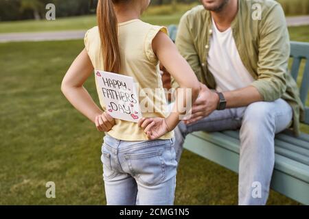 Alles Gute Zum Vatertag. Vater sitzt auf der Holzbank im Park, während seine kleine Tochter versteckt handgemachte Postkarte für ihn Stockfoto