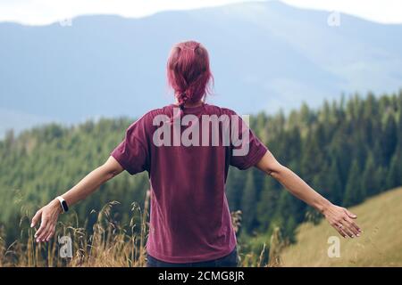 Mädchen mit rosa Haaren auf der Lichtung stehen und schaut auf die Berge. Wandern durch den Wald im Sommer. Dunkler Herbstwald. Lokales Reisekonzept Stockfoto