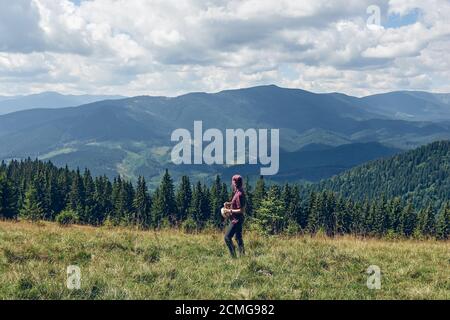 Mädchen mit rosa Haaren auf der Lichtung stehen und schaut auf die Berge. Wandern durch den Wald im Sommer. Dunkler Herbstwald. Lokales Reisekonzept Stockfoto