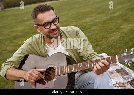 Junger bärtiger Mann mit Brille und Akustikgitarre im Sitzen Auf dem grünen Gras im Park Stockfoto
