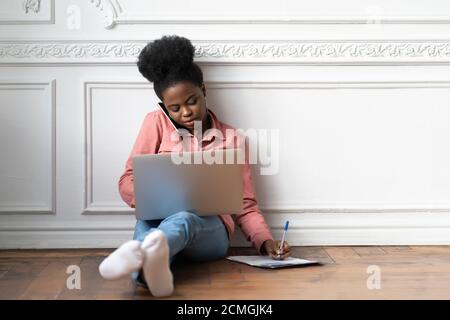 Fokussierte afroamerikanische Frau macht Notizen, klärt Informationen, spricht am Telefon, mit Laptop und sitzen auf dem Boden. Biracial weibliche Mitarbeiter Nachteile Stockfoto