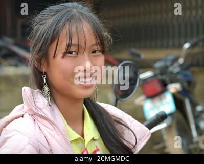 Das moderne vietnamesische Hmong Teenager-Mädchen mit silbernen Ohrringen lächelt für die Kamera vor ihrem Motorrad. Stockfoto