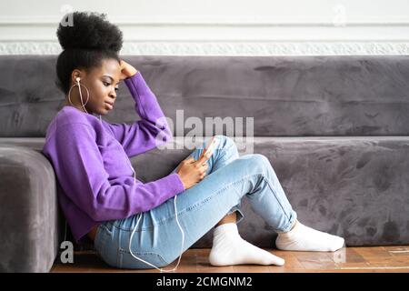 Afroamerikanische Millennial junge Frau in lila Pullover sitzen auf dem Boden, mit Smartphone, Eingabe Nachricht, hört Musik, scrollt durch Stockfoto