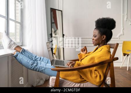 Happy African American Frau Freiberuflerin mit afro Frisur tragen gelbe Strickjacke ruhen, sitzen auf Stuhl, Beine auf Fensterbank lehnend, Remote-Studyin Stockfoto