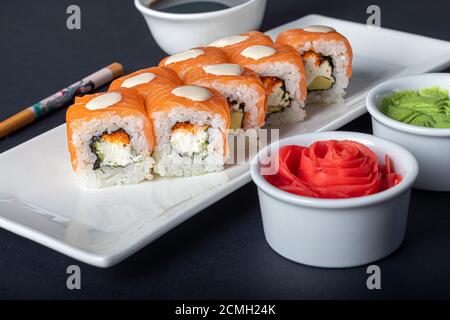 Sushi setzt Uramaki, California, Philadelphia, auf einen weißen Teller. In der Nähe Ingwer und Wasabi. Sojasauce in einer weißen Schüssel. Auf einem dunklen Hintergrund. Speicherplatz kopieren. Stockfoto