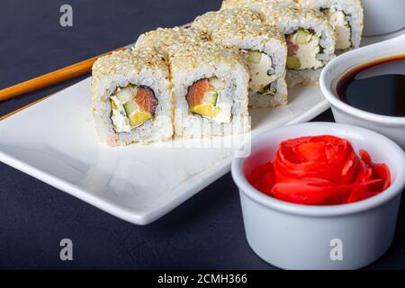 Sushi setzt Uramaki, California, Philadelphia, auf einen weißen Teller. In der Nähe Ingwer und Wasabi. Sojasauce in einer weißen Schüssel. Auf einem dunklen Hintergrund. Speicherplatz kopieren. Stockfoto