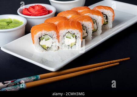Sushi setzt Uramaki, California, Philadelphia, auf einen weißen Teller. In der Nähe Ingwer und Wasabi. Sojasauce in einer weißen Schüssel. Auf einem dunklen Hintergrund. Speicherplatz kopieren. Stockfoto