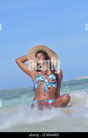 Hispanische Frau sitzt am Sandstrand und lacht mit geschlossenen Augen und Spritzwasser. Los Roques Venezuela Stockfoto