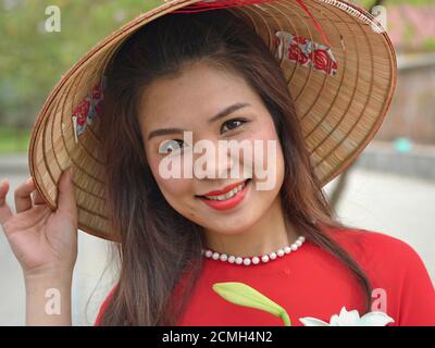 Die junge Vietnamesin in rot trägt einen traditionellen asiatischen konischen Strohhut, hält mit der rechten Hand den Huttrand und posiert für die Kamera. Stockfoto