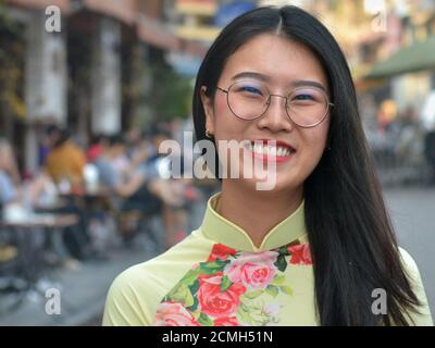 Schöne junge vietnamesische Frau mit langen Haaren trägt moderne Brillen und lächelt für die Kamera vor einem Straßencafé. Stockfoto