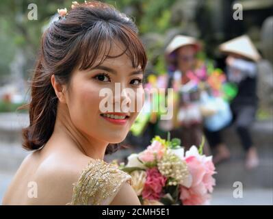 Schöne vietnamesische Braut mit Blumenstrauß Posen für die Kamera, im Hintergrund zwei lokale Frauen mit vietnamesischen konischen Hüten. Stockfoto