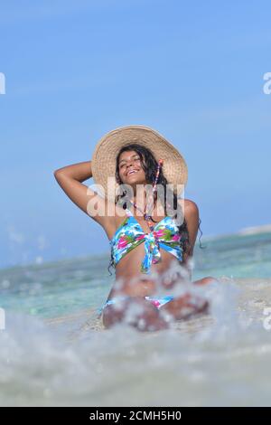 Hispanische Frau sitzt am Sandstrand und lacht mit geschlossenen Augen und Spritzwasser. Los Roques Venezuela Stockfoto