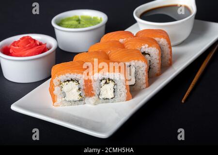 Sushi setzt Uramaki, California, Philadelphia, auf einen weißen Teller. In der Nähe Ingwer und Wasabi. Sojasauce in einer weißen Schüssel. Auf einem dunklen Hintergrund. Speicherplatz kopieren. Stockfoto
