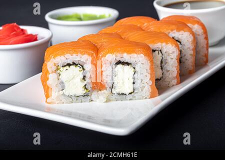 Sushi setzt Uramaki, California, Philadelphia, auf einen weißen Teller. In der Nähe Ingwer und Wasabi. Sojasauce in einer weißen Schüssel. Auf einem dunklen Hintergrund. Speicherplatz kopieren. Stockfoto
