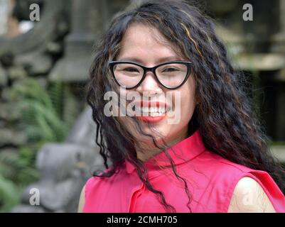 Junge attraktive fröhliche vietnamesische Frau mit großen Brillen trägt eine rosa Bluse und lächelt für die Kamera. Stockfoto