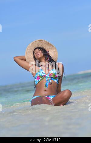 Hispanische Frau sitzt am Sandstrand und lacht mit geschlossenen Augen und Spritzwasser. Glück Stockfoto