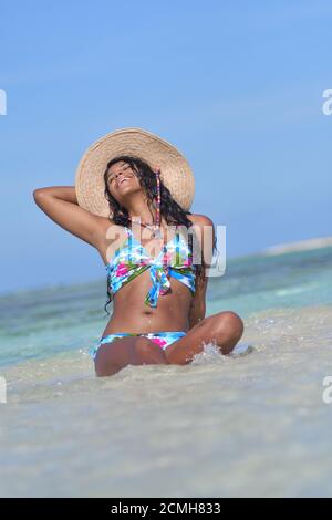 Hispanische Frau sitzt am Sandstrand und lacht mit geschlossenen Augen und Spritzwasser. Glück Stockfoto