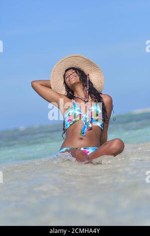 Hispanische Frau sitzt am Sandstrand und lacht mit geschlossenen Augen und Spritzwasser. Glück Stockfoto