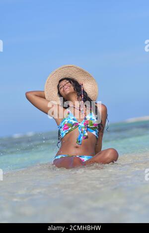 Hispanische Frau sitzt am Sandstrand und lacht mit geschlossenen Augen und Spritzwasser. Glück Stockfoto