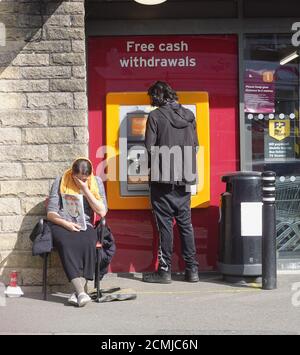 Ein großer Verkäufer außerhalb von Sainsbury's Local und ein Mann am Geldautomaten in New Mills, Derbyshire. Stockfoto