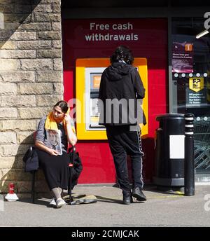 Ein großer Verkäufer außerhalb von Sainsbury's Local und ein Mann am Geldautomaten in New Mills, Derbyshire. Stockfoto