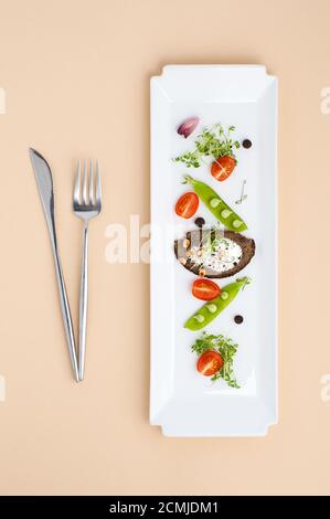 Salat aus jungen grünen Erbsen, Tomaten und Croutons mit Sahne auf einem weißen rechteckigen Teller und Besteck Stockfoto